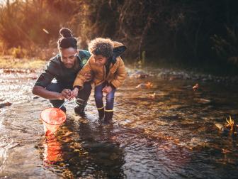 Young dad teaching son how to fish with fishing net in mountain stream at sunset
