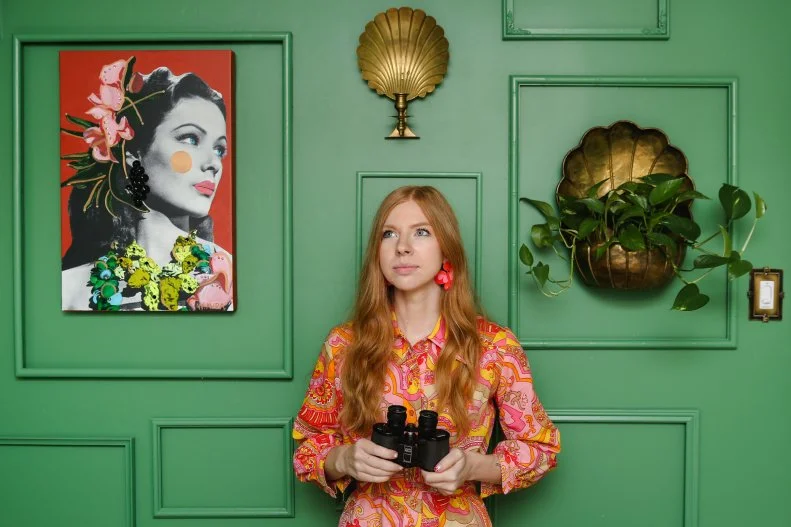 Woman Stands In Front of Green Gallery Wall Holding Binoculars
