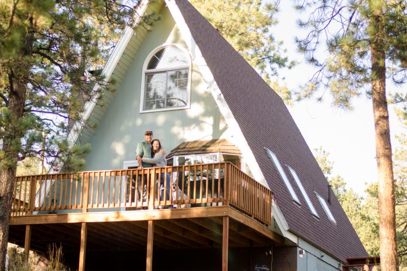 A couple stands on the porch of an A-frame cabin surrounded by pines