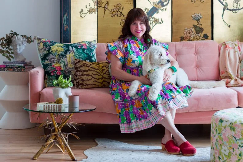 Bari Ackerman sits on a pink couch holding a white Goldendoodle.