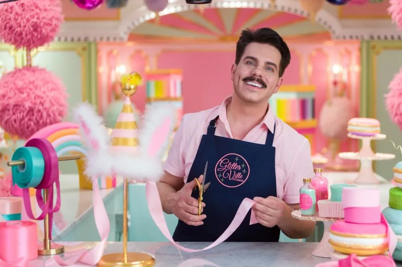 Man smiles to camera in colorful, pink studios holding pink ribbon