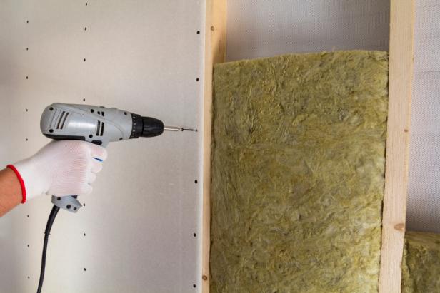 Close-up of worker hand with electric screwdriver fastening drywall to wooden timber frame with insulation mineral rock wool staff. Warm home, low energy bills, construction and renovation concept.