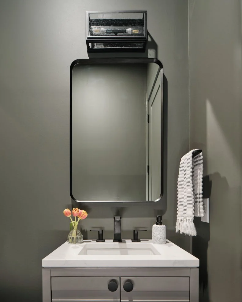 A clean and timeless rectangular mirror with matte black frame sits above the powder room’s grey vanity, with tasteful ship-lap style door panels and coordinating matte black cabinet knobs.