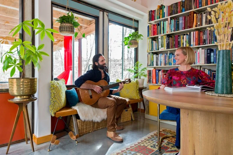 A man with a guitar plays for a woman reading a book at a table.