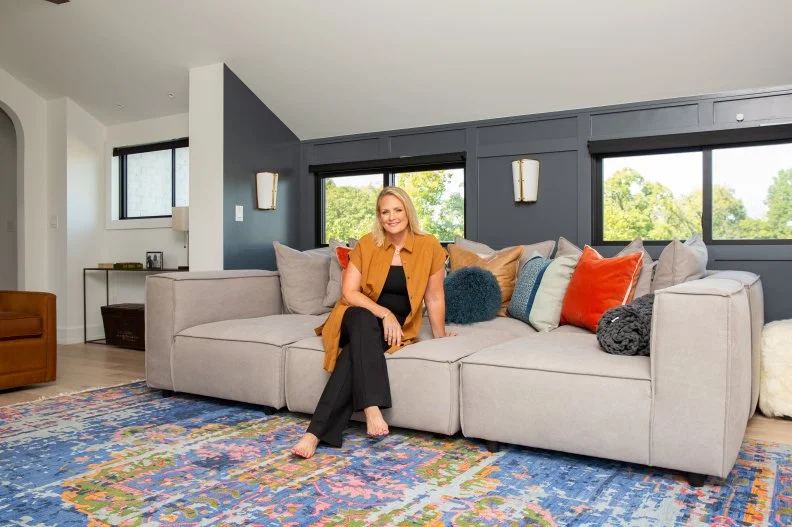 Woman sits on large sofa with pillows in living room, smiles to camera