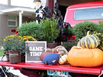Potted mums and shrubs with a basket of apples, pumpkins, footballs and a sign on the open tailgate of a pickup truck for a tailgate party. 