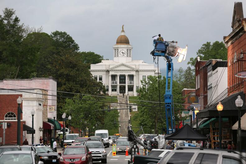 A street view of a movie crew filming "Three Billboards in Ebbing, Missouri, actually set in Sylva, North Carolina, with the county courthouse in the background, cars going by and a movie camera and lights being hoisted into position.