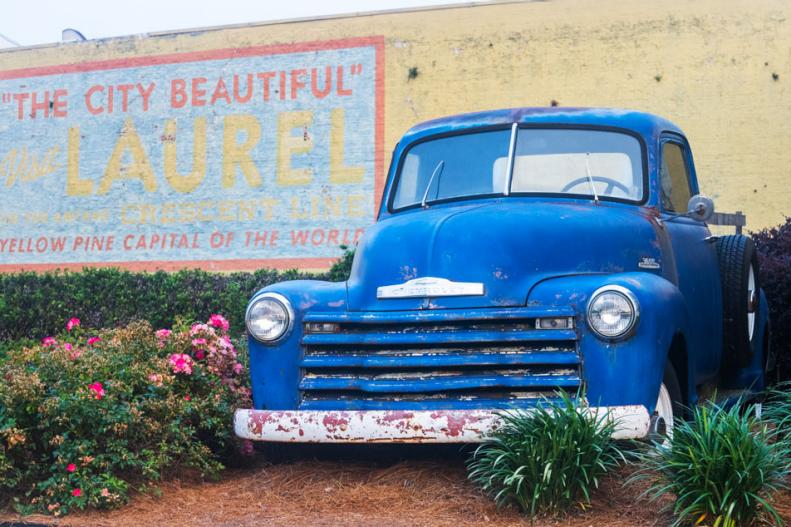 A vintage truck parked in front of a building with a mural about Laurel, Mississippi painted on it.