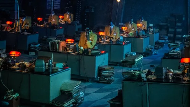 Boiler Room Scene With Desks and Shrinkers from 'Beetlejuice Beetlejuice' Yellow-suited office workers with shrunken heads at matching desks.