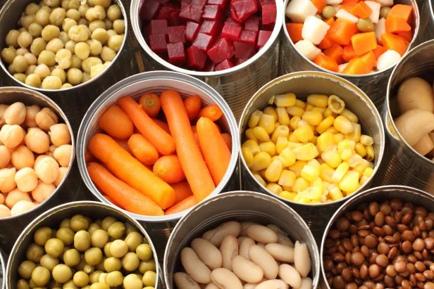 Twelve cans of open vegetables sit next to one another in three rows.  There are peas, carrots, beats, snow peas, corn, chick peas and Lima beans in the cans.