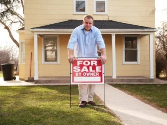 Photo of a young man posting a "for sale by owner" sign in the front yard of a very tiny yellow house.