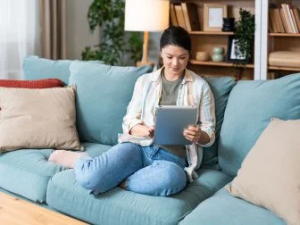 Young design professional business woman sitting on sofa at home using digital tablet. Female choosing new wardrobe from internet store for online shopping and ordering.