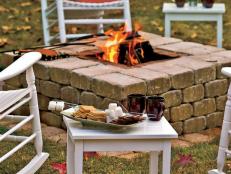 A roaring fire pit is surrounded with chairs and a camping-style plate of  s'mores.