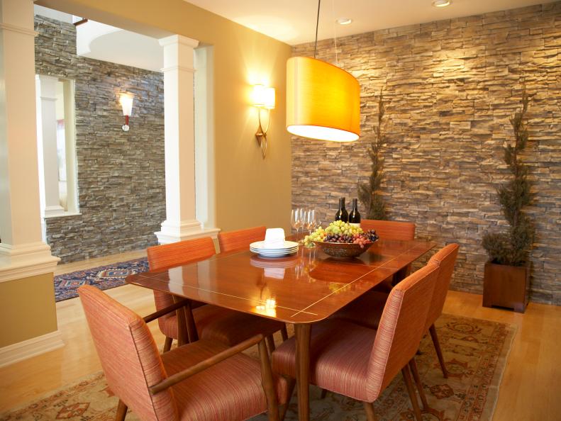 a stone wall makes a dramatic statement in this contemporary dining space.hardwood floors are warmed with an area rug that coordinates with the orange and red contemporary furnishings.a unique lighting fixture hangs over the dining table and cast warmth. 