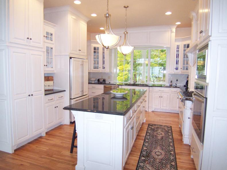 Wide view of white kitchen with light hardwood flooring, rug, appliances, kitchen island with new countertop, lighting, and white cabinetry. 
