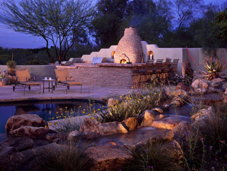Outdoor entertaining area with grill and stone fireplace, bar area with chairs, pond with plants, rocks, and flowers, nighttime view of outdoor room. 