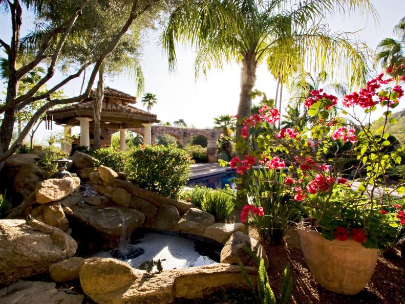 Tropical landscape design with pink flowers in a pot, water feature, gazebo with columns, and several plants and decor. 