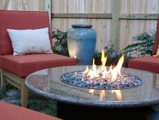 As seen on Indoors Out, this is a closeup of a fire pit insert with a burning flame in an outdoor space with outdoor furniture and a privacy wood wall. 