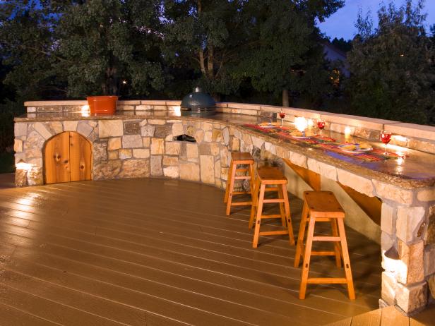 Outdoor Kitchen at dusk with wood barstools, stone frame, and wood deck. 