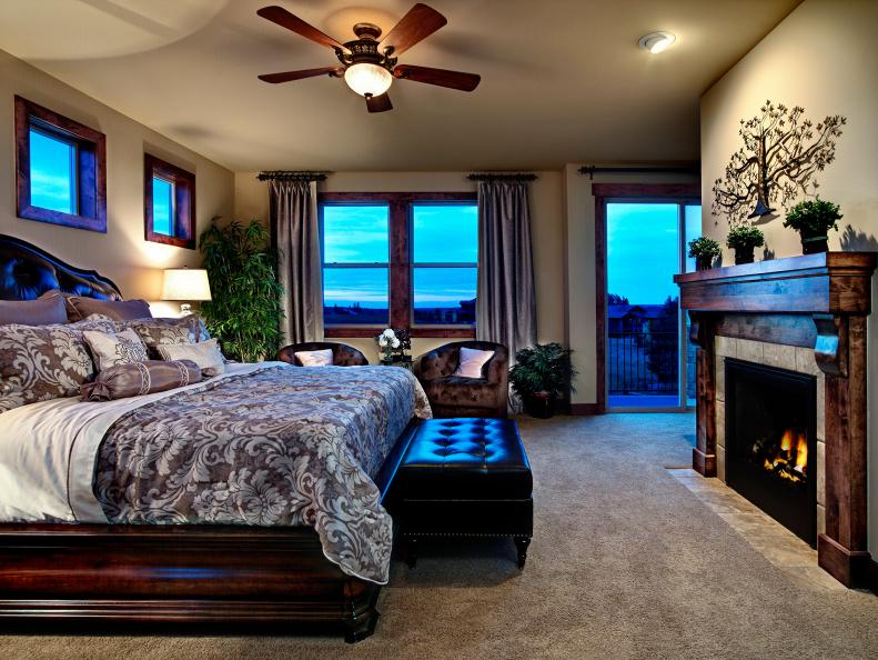 Bedroom side view with fireplace and mantel, tree art on wall, warm wall tones, ceiling fan, doors leading outside, black bench at the foot of the bed, and sitting area. 