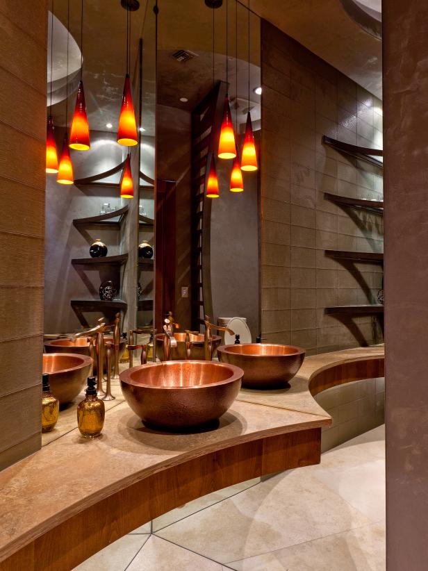 This contemporary southwestern bathroom has a dramatically curved quartz slab top with a wooden apron that comes to a point and copper sinks and bronze fixtures on top. The sculptural open shelving is curved too. The rustic textures, neutral colors, the mix of wood and stone brings warmth to the space. The bathroom has simple lighting by LED rope lighting concealed under the counter, ceiling soffit, and hanging pendants.