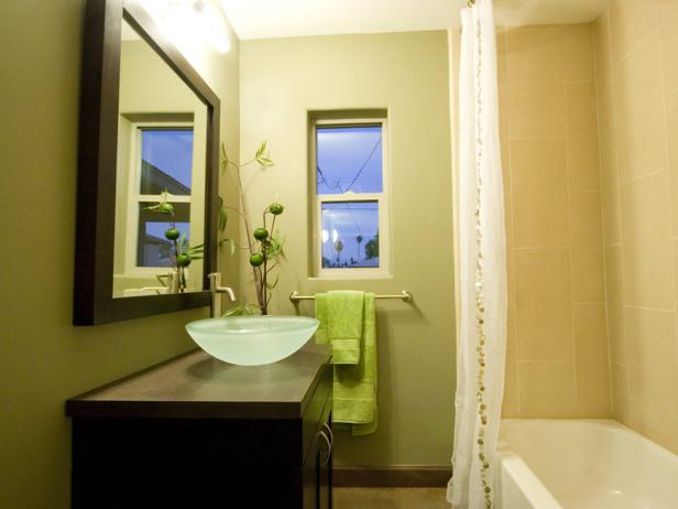 Side view of a green and brown bathroom design, vanity, large mirror, glass sink bowl, shower, tub, and pale green walls, and towel holder. 