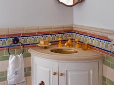 A corner of this Mediterranean-inspired bathroom includes a large mirror, hung at an angle; a wedge-shaped vanity with round inset sink, gold fixtures, and space for soap dish and candleholder; and a tile wall with alternating rows of green and white tile which is topped by a row of blue, yellow, and green tiles that form a pastoral scene.