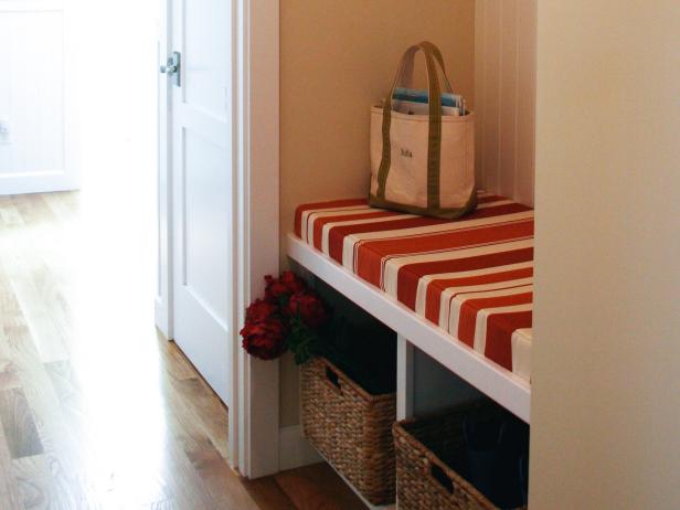 A padded white bench is used in this mudroom for storage. Beneath the bench are two wicker baskets, while above the bench is an area to hang coats and additional open-faced cabinets.