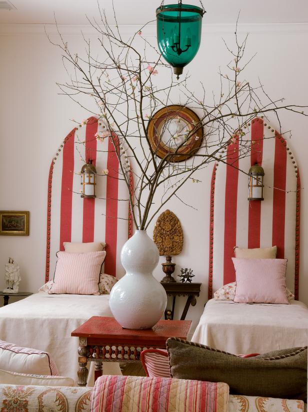 This bedroom has cinnabar red accents and two twin beds. It has a Gothic charm, with the walls painted in clean white . Red and white striped vertical upholstered boards serve as the headboard in the room. 
