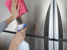 The woman cleaning surface of inox refrigerator with pink rag and spray. 