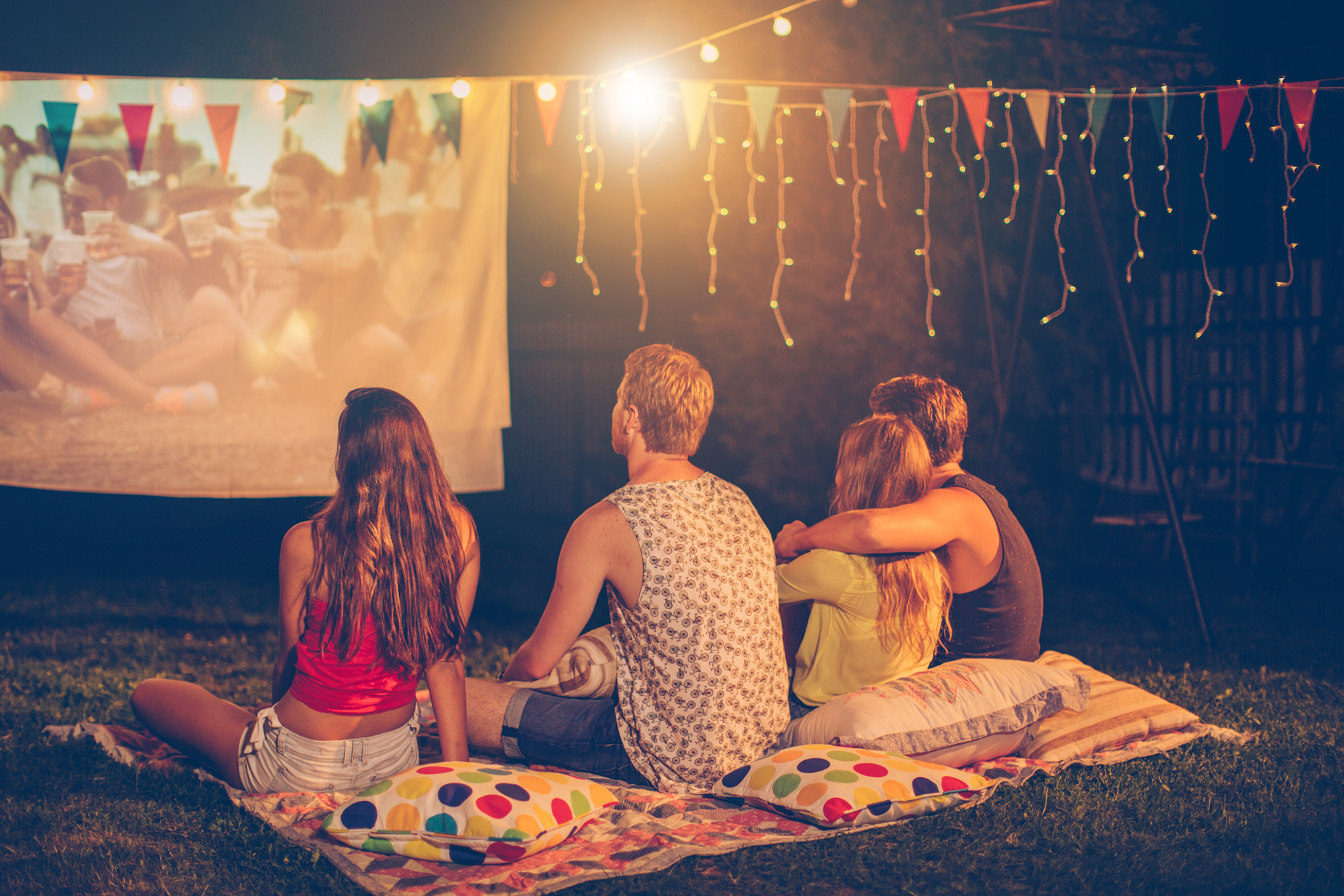 Young friends having movie night party. Laying down on blanket in front of movie improvised screen. Backyard decorated with festive string lights. Night time. View from behind.