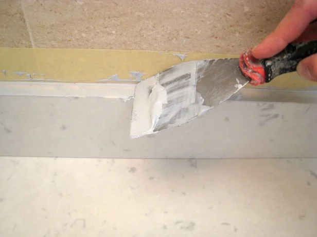 Apply caulk with a putty knife, pressing down into the seams, then pulling the knife along the length of the backsplash to smooth out the caulk.