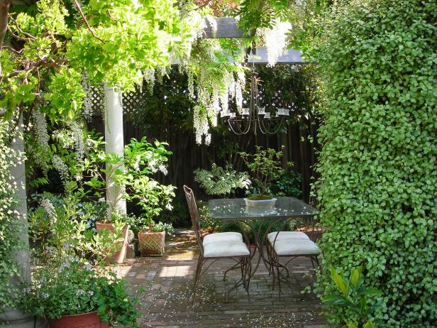 A glass table and metal chairs are surrounded by abundant garden landscaping in this outdoor patio dining area. A candle chandelier hangs from an overhead pergola.