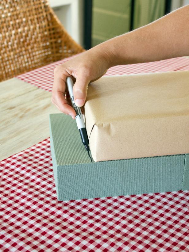 To create a gift box centerpiece, place a wrapped box on top of (and in the center of) a larger floral foam gift box, and use a pen or marker to trace a line onto the floral foam at each end of the box.