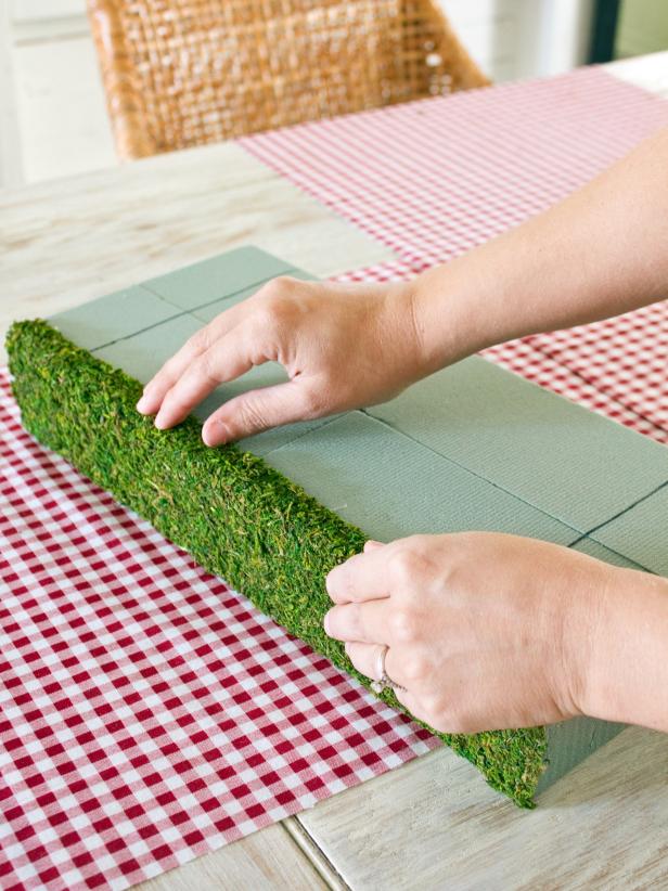 To create a moss-covered gift box centerpiece, use scissors to cut out a piece of sheet moss the same size as your floral foam. Attach it to the front of the floral foam gift box with hot glue. Continue to attach moss to each side until the box is covered (except for the area where the center box will sit).