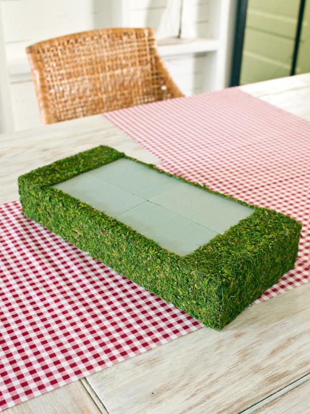 When making a moss-covered gift box centerpiece, use scissors to cut out a piece of sheet moss the same size as your floral foam, and attach it to the front of the floral foam gift box with hot glue. Continue to attach moss to each side until the box is covered (except for the area where the center box will sit).