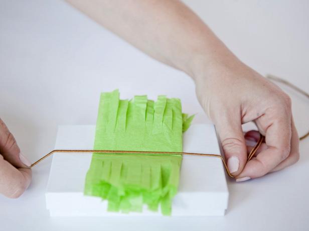 Unfold the fringed tissue paper once and tie onto a gift box with a gold metallic cord. Knot the cord on the back and cut off any extra. Turn the box over and fluff and crunch the fringed tissue paper to create a fun pouf.