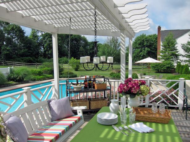 White furniture under a white pergola gives a fresh look to this outdoor spot for afternoon tea. A hanging chandelier with white votive candles and a hydrangea arrangement bring additional dimension to the space, which sits by a relaxing swimming pool and gorgeous landscape.