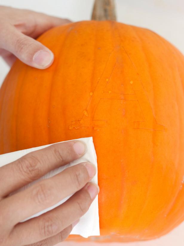 Using an X-Acto knife, trace the letter's outline onto the pumpkin. Tip: If the letter has any inside lines, cut those out first. Use a napkin or paper towel to wipe off any juice that may have seeped out during the tracing process. To produce a monogrammed silhouette of the letter A on this Halloween pumpkin, one must first wipe the pumpkin with water.