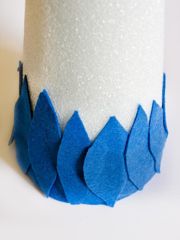 Apply hot glue in small sections around the base of each tree form and immediately press strip of darkest felt in place. Just above felt strip, apply a bead of hot glue vertically and immediately press the darkest felt feather into glue. Hold in place for few seconds until glue cools. Repeat with other dark felt feathers, placing them side by side so they slightly overlap, concealing the cone. Once the entire bottom row is finished, start a second row of dark felt feathers just above first row. Continue to glue feathers on, graduating from dark to light in rows of two. Tip: Use a moderate amount of hot glue. You don't want glue to squeeze out when felt feathers are pressed down.