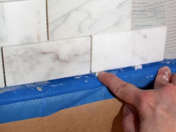 As you apply sheets of tile, slide spacers under the bottom row of tile to create a gap that later will be filled with caulk. It's better to caulk, rather than grout, this bottom line to allow for cabinets shifting with normal floorboard movement.