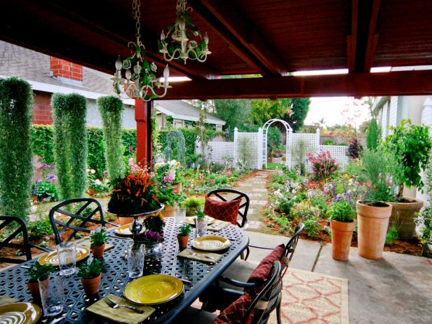 This beautifully landscaped courtyard includes an arched arbor and stone pathway that leads to a covered patio and outdoor dining area surrounded by container plants and tall topiaries. Traditional chandeliers are covered in leafy vines to continue the garden look.