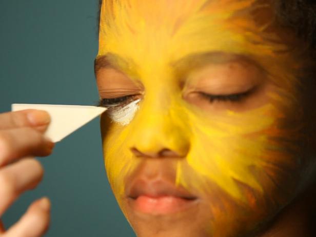 To create a base for your lion makeup, begin by dampening a sponge then picking up some of the yellow face paint. Tip: When using dry face paints, be sure to get sponge or brush wet before dipping into paint. Have child close their eyes, then apply yellow paint to the center of their face and blend out. Dip a clean sponge in water then in white paint and apply under eye, angling down the sides of the nose. Add white above top lip, blending out onto cheeks and down chin to resemble a lion's muzzle.