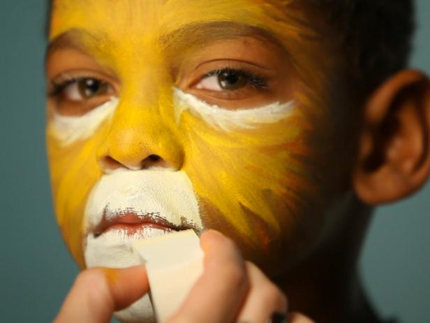 To create a base for your lion makeup, begin by dampening a sponge then picking up some of the yellow face paint. Tip: When using dry face paints, be sure to get sponge or brush wet before dipping into paint. Have child close their eyes, then apply yellow paint to the center of their face and blend out. Using the same sponge, add orange to the yellow paint's edges and blend out further to create a blended ombre effect. Apply orange paint like brushstrokes to resemble fur. Dip a clean sponge in water then in white paint and apply under eye, angling down the sides of the nose. Add white above top lip, blending out onto cheeks and down chin to resemble a lion's muzzle.