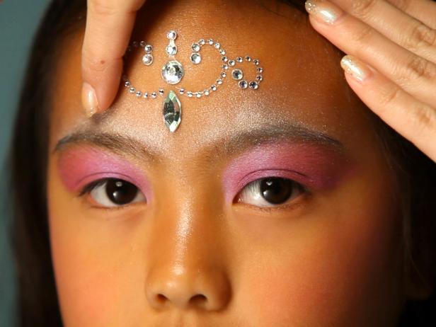 To apply fairy princess makeup, begin with a pearly eye shadow. Then add pink blush on the cheeks. Next, apply self-adhesive glittering jewels on forehead like a princess' tiara, and then apply additional jewels under eyebrows, on cheekbones or anywhere you like. Tip: You can find self-adhesive jewels in the scrapbooking section of your local craft store. Make sure your child doesn't have any adhesive allergies before continuing with this step. Keeping away from the sensitive eye area, dab face lotion on the cheeks and surrounding the jewel tiara on forehead. Have model close her eyes then apply cosmetic glitter where lotion was applied. Paint lips with a sparkly pink lipstick or tinted lip-gloss. Finish the look with a fairy princess costume.