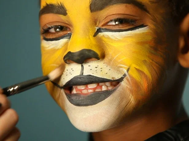 To give your child a ferocious lion’s face for Halloween, simply paint a basic orange and yellow face. Then paint their top lip black and extend curving line up cheeks slightly. Paint their bottom lip black, then add black dots to white area above top lip. Add whiskers by brushing white paint in small, short strokes around mouth and cheek area. Add a line of black paint under eyes, angling down the nose to create those striking big-cat eyes. Use a blending brush and brown eye shadow to shade down sides of nose, down the smile crease and chin, and create some brush texture around edges of face to look like fur. Add a few strokes of white to the forehead and chin for added depth and definition.