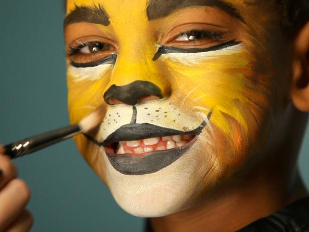 To give your child a ferocious lion’s face for Halloween, simply paint a basic orange and yellow face. Then paint their top lip black and extend curving line up cheeks slightly. Paint their bottom lip black, then add black dots to white area above top lip. Add whiskers by brushing white paint in small, short strokes around mouth and cheek area. Add a line of black paint under eyes, angling down the nose to create those striking big-cat eyes. Use a blending brush and brown eye shadow to shade down sides of nose, down the smile crease and chin, and create some brush texture around edges of face to look like fur. Add a few strokes of white to the forehead and chin for added depth and definition.