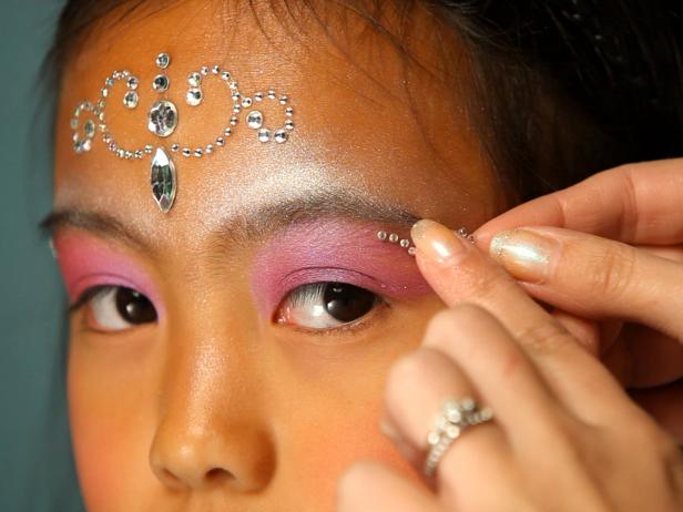 To apply fairy princess makeup, begin with a pearly eye shadow. Then add pink blush on the cheeks. Next, apply self-adhesive glittering jewels on forehead like a princess' tiara, and then apply additional jewels under eyebrows, on cheekbones or anywhere you like. Tip: You can find self-adhesive jewels in the scrapbooking section of your local craft store. Make sure your child doesn't have any adhesive allergies before continuing with this step. Keeping away from the sensitive eye area, dab face lotion on the cheeks and surrounding the jewel tiara on forehead. Have model close her eyes then apply cosmetic glitter where lotion was applied. Paint lips with a sparkly pink lipstick or tinted lip-gloss. Finish the look with a fairy princess costume.