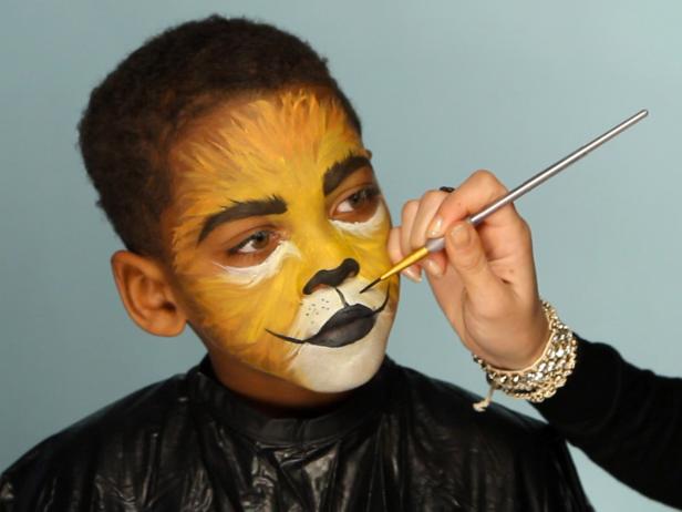 To give your child a ferocious lion’s face for Halloween, simply paint a basic orange and yellow face. Then paint their top lip black and extend curving line up cheeks slightly. Paint their bottom lip black, then add black dots to white area above top lip. Add whiskers by brushing white paint in small, short strokes around mouth and cheek area.
