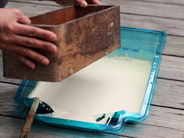 Step 3 in crafting a chic bathroom caddy is to dip the bottom of a wooden box into a pan of paint. Repeat several times until you get the perfect amount of paint on the bottom of the box. Allow the box to dry.